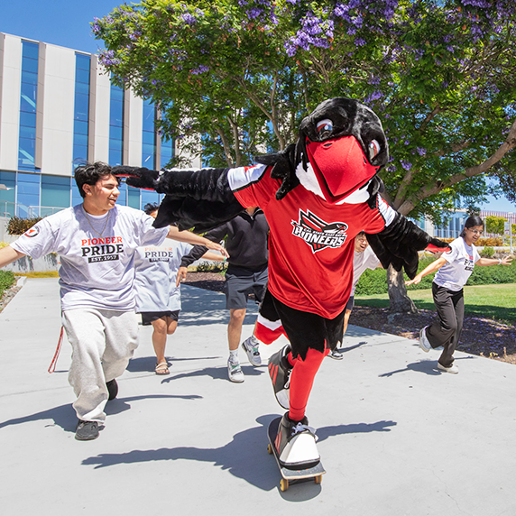 Cal State East Bay’s mascot Perry the Peregrine Falcon skateboards down a campus walkway with students running and smiling behind them.