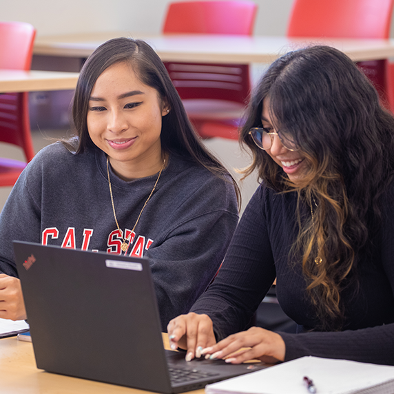 Two students sit together in a classroom, smiling as they work on a laptop and take notes.