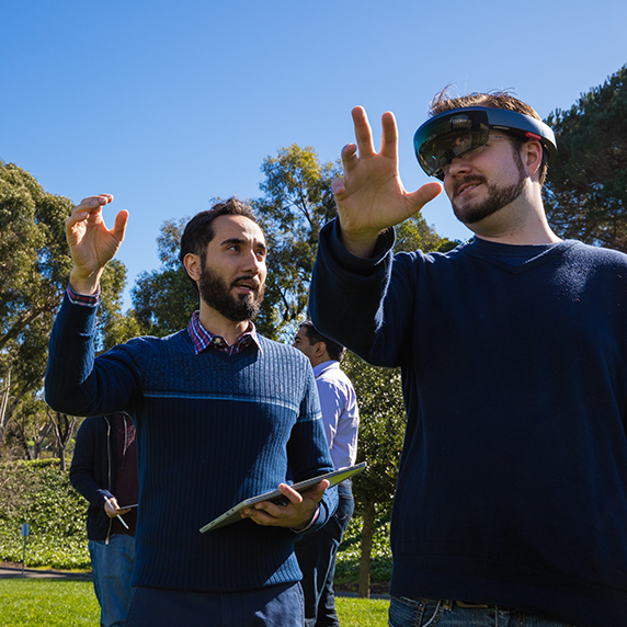 Two students stand outdoors on a sunny day, one holding a tablet and the other wearing an augmented-reality headset, both raising their hands as they interact with the technology.